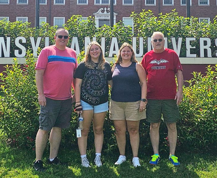 four people posing in front of a campus sign