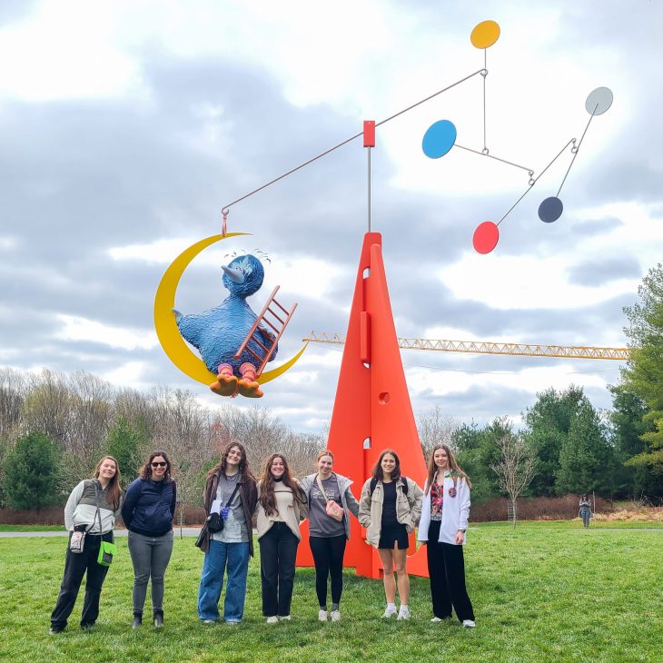 Seven students pose in front of a large outdoor sculpture featuring a blue bird sitting on a crescent moon