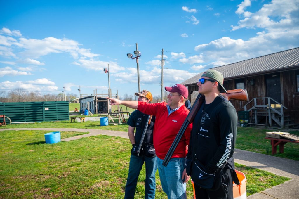 three men looking out into a field man in the center is wearing a read sweater and pointing at something