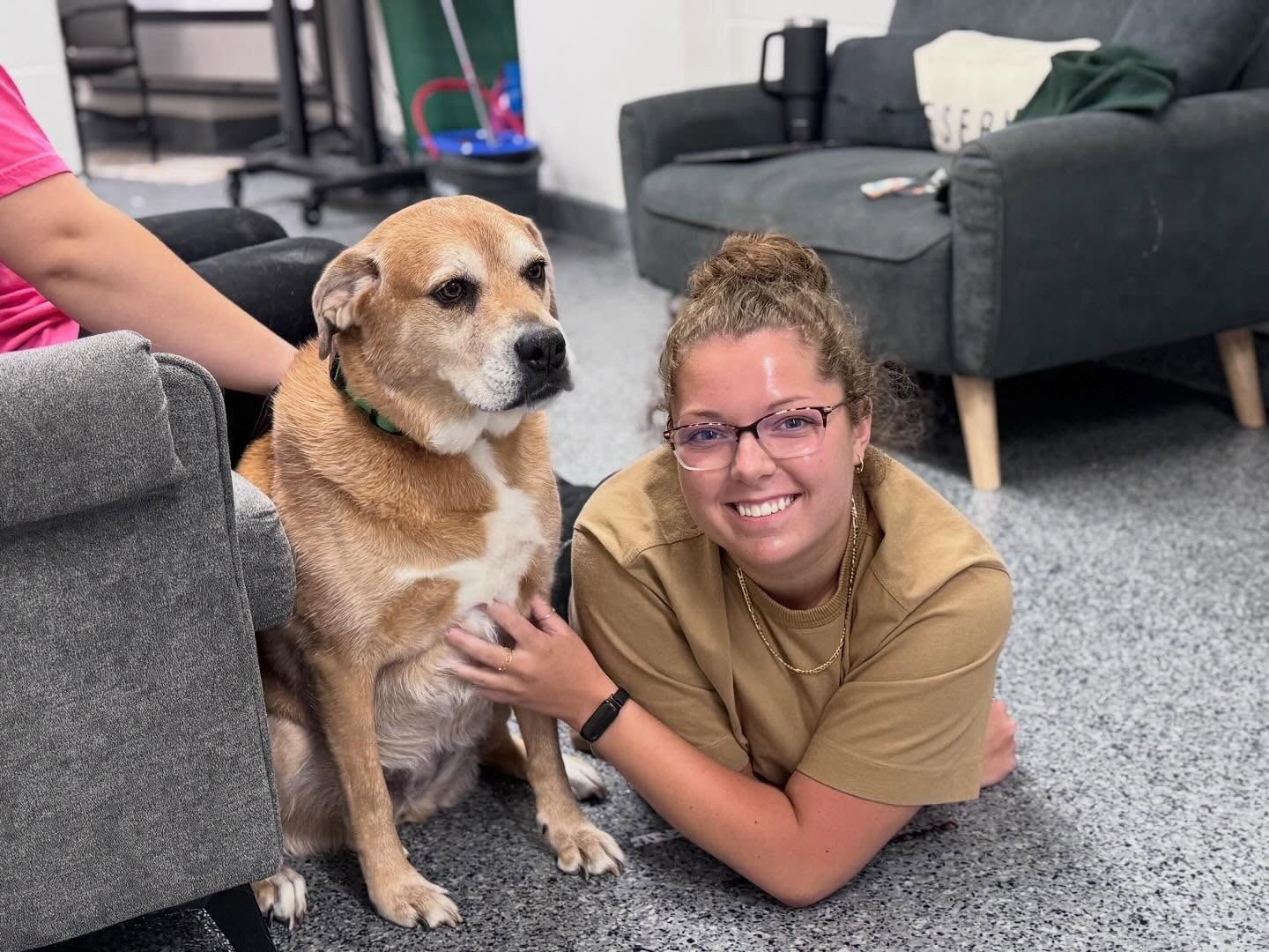 A smiling woman with glasses lying on the floor beside a seated tan dog, touching its chest