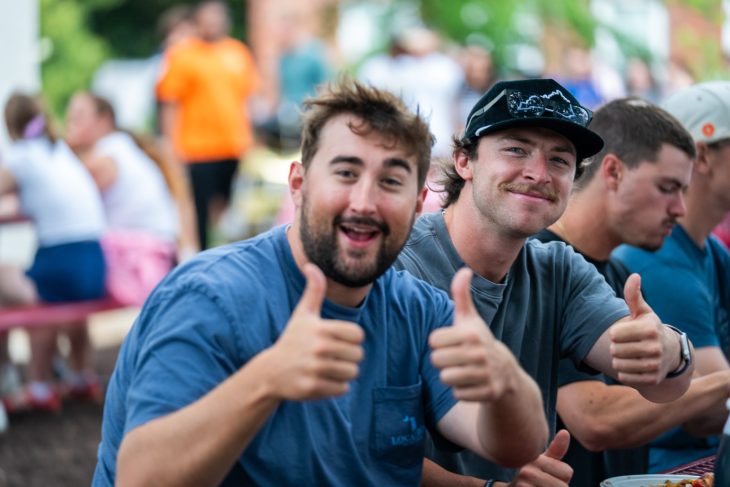 Two smiling students give thumbs up while sitting outdoors on campus
