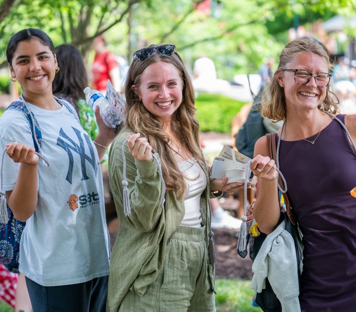 Three smiling students holding white academic honor cords outdoors