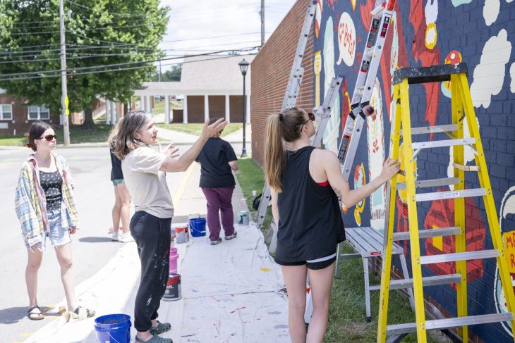 students and professor gathered around the mural in progress with painting supplies and ladders