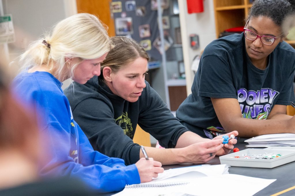 Three students collaborate on a science project using a molecular model kit in a classroom